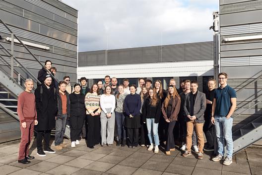 You see a group of about 20 students in a group photo. They are standing on a terrace of the buildings at Hochschule Düsseldorf. In the background, the well-known silver-metal facade is visible.